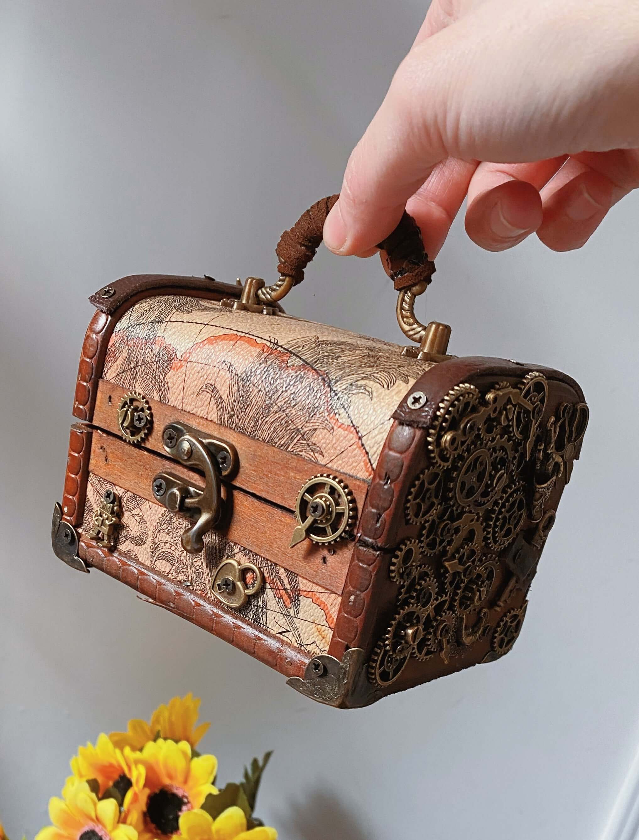 Handmade steampunk jewelry storage box with gears and a decorative clasp, held in a hand with sunflowers in the background.